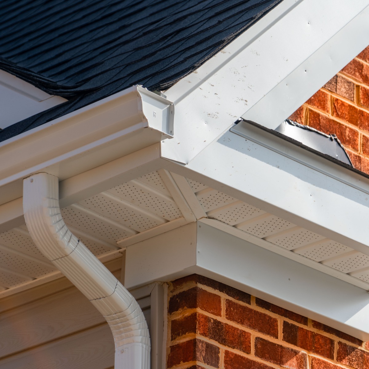 Closeup view of white gutter system with soffit vent, gutter guard, drop outlet, downspout, vinyl elbows, decorative trim molding, on the corner of a brick luxury house in America